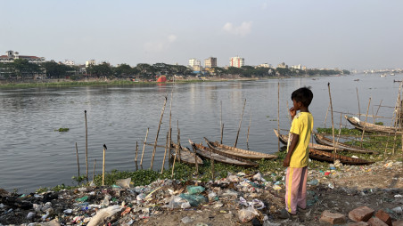 A child, wearing a bright yellow t-shirt, stands atop garbage with dinghy boats anchored near the Buriganga River, while looking at the cityscape on the other side. Photo: Yasin Piash/TBS 
