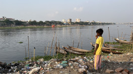A child, wearing a bright yellow t-shirt, stands atop garbage with dinghy boats anchored near the Buriganga River, while looking at the cityscape on the other side. Photo: Yasin Piash/TBS 